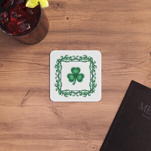 May include: Square white coaster featuring a green shamrock design within a green decorative border. The coaster sits on a wooden surface, with a glass of red liquid and a menu in the background.
