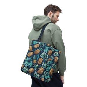 May include: A tote bag with a repeating pattern of pineapples and tropical leaves in shades of gold, teal, and navy blue. The bag has a navy blue strap and is carried over the shoulder.