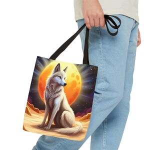 Könnte beinhalten: Ein weißer Wolf mit blauen Augen sitzt vor einem großen orangefarbenen Mond. Der Wolf ist von einem sternenklaren Nachthimmel umgeben. Das Bild ist auf einer Tragetasche gedruckt.