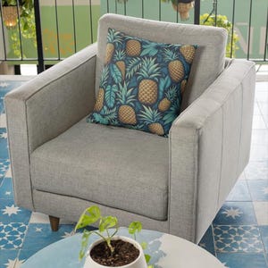 May include: A gray armchair with a decorative pillow featuring a pineapple pattern in shades of gold, green, and blue. The chair is set in a room with a blue and white patterned floor and a small white pot with a plant on a round table.