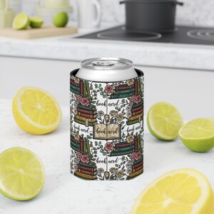 May include: A white can cooler featuring a repeating pattern of stacked books, roses, and the words "book nerd." The design includes green, pink, and brown tones. The cooler is designed to keep beverages cold.