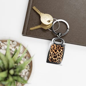 Può includere: Un portachiavi d'argento con un ciondolo rettangolare con un design a stampa leopardata. Il portachiavi ha un anello d'argento e un anello d'argento.