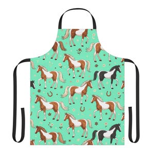 May include: A kitchen apron featuring a repeating pattern of brown and white horses, black and white horses, horseshoes, and small flowers on a mint green background. The apron includes black neck and waist ties for securing the garment.
