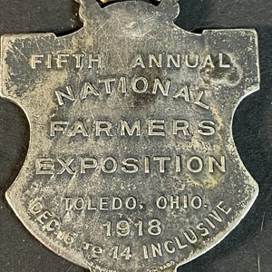 Puede incluir: Una medalla plateada vintage con forma de escudo y una correa de cuero desgastada. El texto dice "FIFTH ANNUAL NATIONAL FARMERS EXPOSITION" y "TOLEDO, OHIO. DEC 6 TO 14 INCLUSIVE 1918".