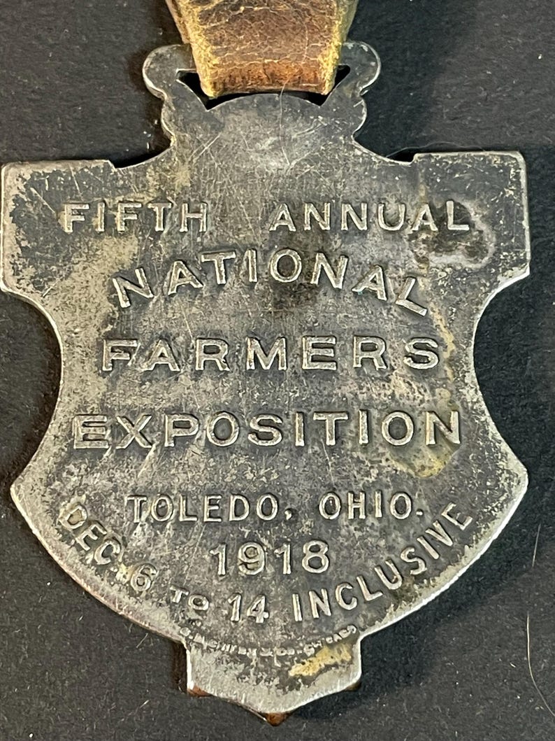 Puede incluir: Una medalla plateada vintage con forma de escudo y una correa de cuero desgastada. El texto dice "FIFTH ANNUAL NATIONAL FARMERS EXPOSITION" y "TOLEDO, OHIO. DEC 6 TO 14 INCLUSIVE 1918".