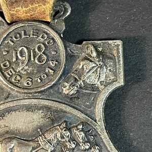 Puede incluir: Medalla plateada con correa de cuero marr&oacute;n. La medalla presenta el texto "TOLEDO 1918" y una representaci&oacute;n de caballos. La medalla tiene un dise&ntilde;o detallado con una est&eacute;tica vintage.