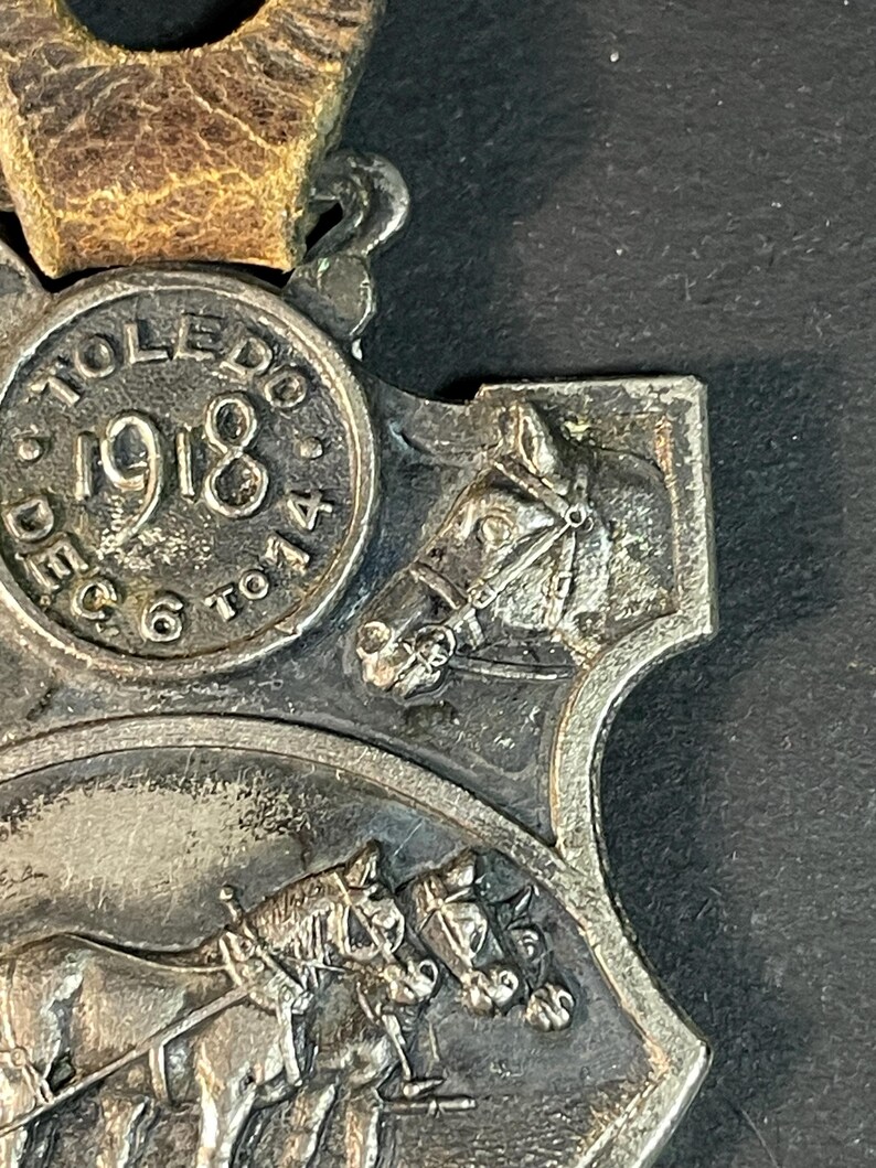 Puede incluir: Medalla plateada con correa de cuero marr&oacute;n. La medalla presenta el texto "TOLEDO 1918" y una representaci&oacute;n de caballos. La medalla tiene un dise&ntilde;o detallado con una est&eacute;tica vintage.