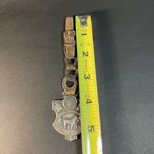 Puede incluir: Un llavero vintage de metal plateado y cuero con un dise&ntilde;o de escudo. El llavero presenta una escena con caballos y un carruaje, y el a&ntilde;o 1918. La correa de cuero mide aproximadamente 5 cm de largo.