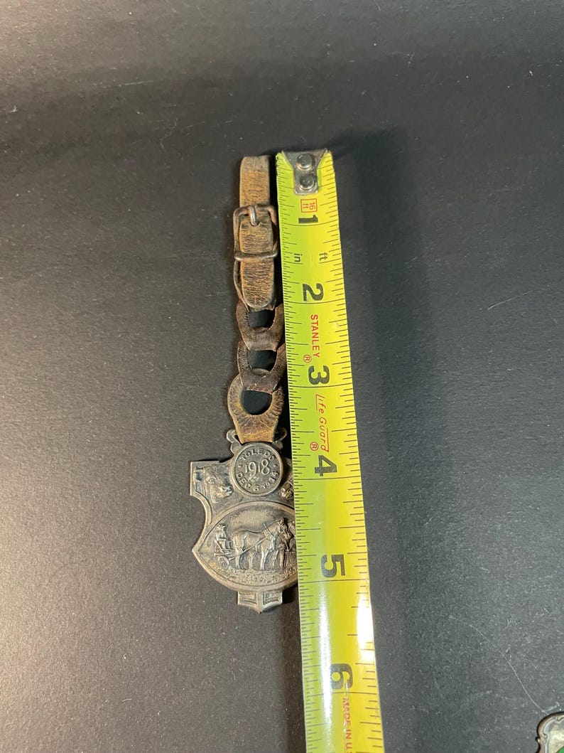 Puede incluir: Un llavero vintage de metal plateado y cuero con un dise&ntilde;o de escudo. El llavero presenta una escena con caballos y un carruaje, y el a&ntilde;o 1918. La correa de cuero mide aproximadamente 5 cm de largo.