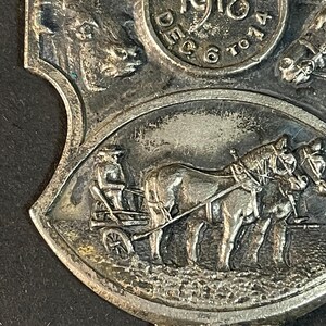 Puede incluir: Medalla antigua de color plateado con detalles intrincados. La medalla presenta el a&ntilde;o "1918" y la palabra "Toledo" en un dise&ntilde;o circular. Debajo, se representa un carruaje tirado por caballos. La medalla tiene forma de escudo.