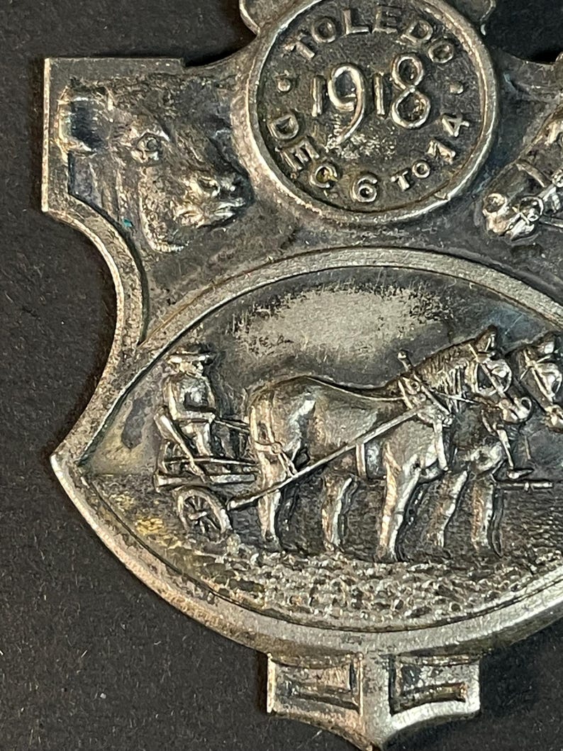 Puede incluir: Medalla antigua de color plateado con detalles intrincados. La medalla presenta el a&ntilde;o "1918" y la palabra "Toledo" en un dise&ntilde;o circular. Debajo, se representa un carruaje tirado por caballos. La medalla tiene forma de escudo.