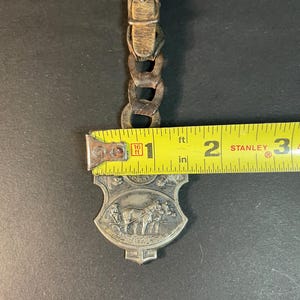 Puede incluir: Un llavero vintage de metal plateado con un carruaje tirado por caballos, con una correa y hebilla de cuero. El llavero mide aproximadamente 5 cm de largo y presenta un relieve detallado de un carruaje tirado por caballos. La correa es de color marr&oacute;n claro.