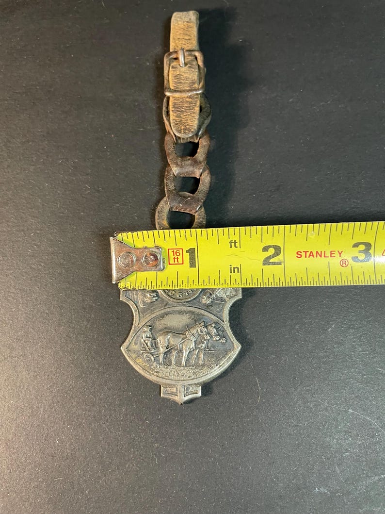 Puede incluir: Un llavero vintage de metal plateado con un carruaje tirado por caballos, con una correa y hebilla de cuero. El llavero mide aproximadamente 5 cm de largo y presenta un relieve detallado de un carruaje tirado por caballos. La correa es de color marr&oacute;n claro.