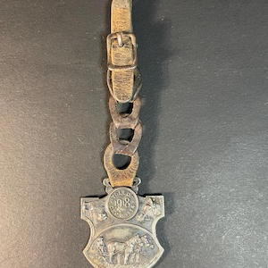 Puede incluir: Un guardatiempos de bolsillo antiguo de tono plateado con correa y cadena de cuero. El guardatiempos presenta un dise&ntilde;o de escudo con el a&ntilde;o "1918" y una representaci&oacute;n de caballos tirando de un carruaje. La correa de cuero tiene una hebilla.