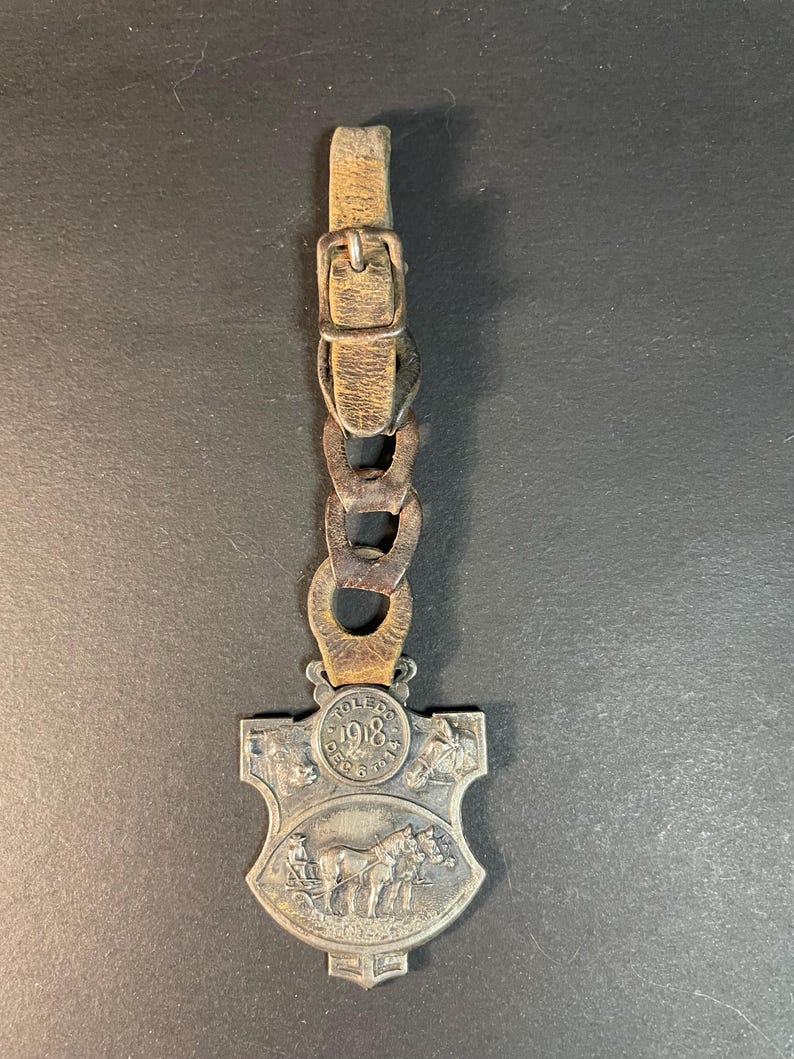 Puede incluir: Un guardatiempos de bolsillo antiguo de tono plateado con correa y cadena de cuero. El guardatiempos presenta un dise&ntilde;o de escudo con el a&ntilde;o "1918" y una representaci&oacute;n de caballos tirando de un carruaje. La correa de cuero tiene una hebilla.