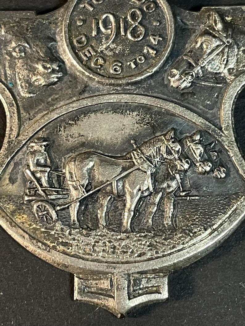 Puede incluir: Primer plano de una medalla de metal plateado con im&aacute;genes en relieve. El dise&ntilde;o central presenta un carruaje tirado por caballos con un conductor. La parte superior tiene el a&ntilde;o "1918" y las palabras "Dec 6 to 14". La medalla tiene una est&eacute;tica vintage detallada.
