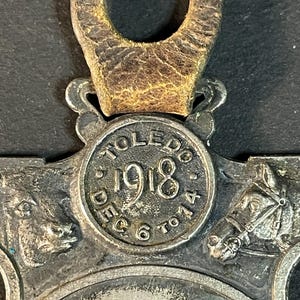 Puede incluir: Primer plano de un medall&oacute;n vintage de metal plateado con una correa de cuero. El medall&oacute;n presenta el texto "TOLEDO 1918 DEC 6 TO 14" e im&aacute;genes en relieve detalladas de una vaca, un caballo y un carruaje tirado por caballos. La correa de cuero es marr&oacute;n.