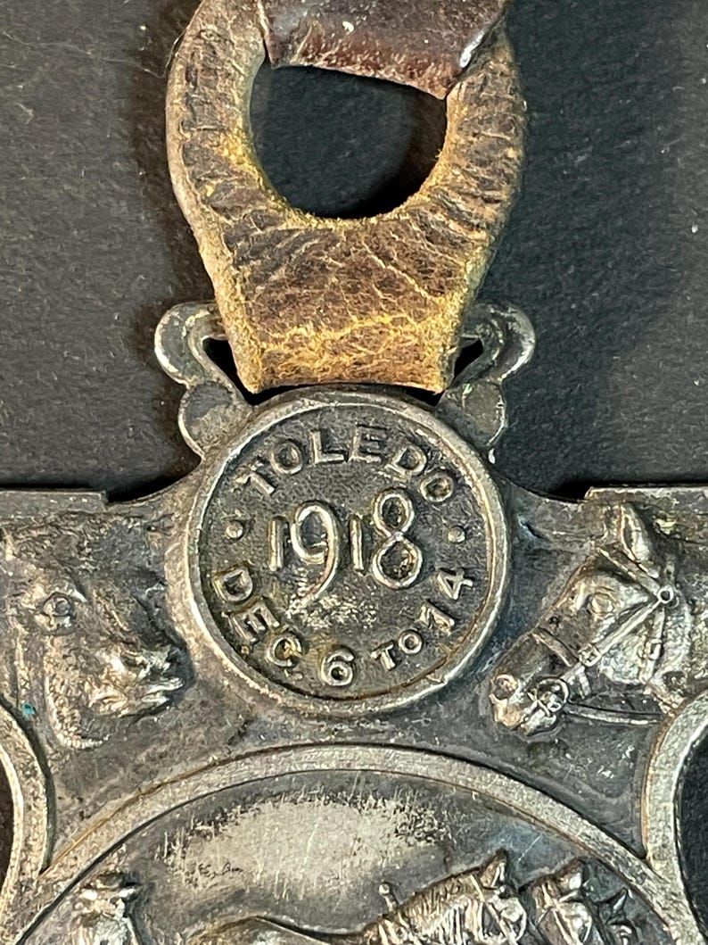 Puede incluir: Primer plano de un medall&oacute;n vintage de metal plateado con una correa de cuero. El medall&oacute;n presenta el texto "TOLEDO 1918 DEC 6 TO 14" e im&aacute;genes en relieve detalladas de una vaca, un caballo y un carruaje tirado por caballos. La correa de cuero es marr&oacute;n.
