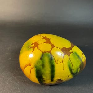 May include: A decorative, egg-shaped object with a glossy finish. The egg features a vibrant color palette of yellow, brown, and green, with a unique, abstract pattern. The design includes brown, web-like patterns over a yellow base, and green streaks.