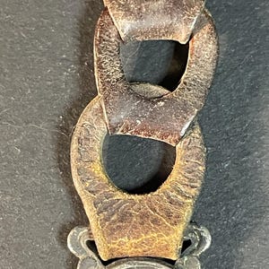 Puede incluir: Primer plano de una cadena met&aacute;lica vintage con eslabones grandes y ovalados. La cadena es de color marr&oacute;n oscuro y desgastado, mostrando signos de edad y desgaste. La cadena est&aacute; conectada a un emblema plateado con la palabra "TOLEDO" visible.