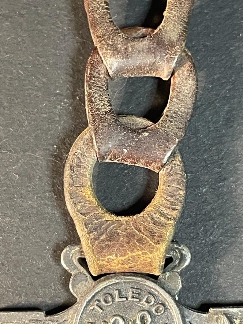 Puede incluir: Primer plano de una cadena met&aacute;lica vintage con eslabones grandes y ovalados. La cadena es de color marr&oacute;n oscuro y desgastado, mostrando signos de edad y desgaste. La cadena est&aacute; conectada a un emblema plateado con la palabra "TOLEDO" visible.