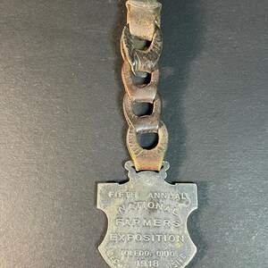 Puede incluir: Una medalla vintage de metal en forma de escudo con el texto "FIFTH ANNUAL NATIONAL FARMERS EXPOSITION TOLEDO, OHIO 1918 DEC 9-14 INCLUSIVE" adjunta a una correa de cuero con eslabones de cadena de metal. La medalla tiene un acabado plateado deslustrado.