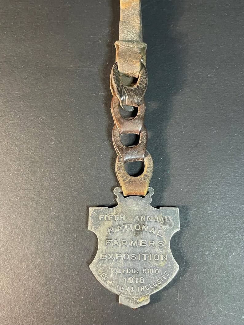 Puede incluir: Una medalla vintage de metal en forma de escudo con el texto "FIFTH ANNUAL NATIONAL FARMERS EXPOSITION TOLEDO, OHIO 1918 DEC 9-14 INCLUSIVE" adjunta a una correa de cuero con eslabones de cadena de metal. La medalla tiene un acabado plateado deslustrado.