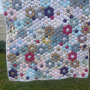 Könnte beinhalten: Eine handgefertigte Steppdecke mit floralem Muster in verschiedenen Farben wie Rosa, Blau, Lila und Weiß. Die Steppdecke besteht aus sechseckigen Stoffstücken, die ein einzigartiges Design ergeben. Die Steppdecke hängt draußen an einer Wäscheleine.