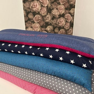 May include: A stack of six long, rectangular pillows with different patterns. The top pillow is blue with white stars and the text "Fantasie Baby" embroidered in pink. The other pillows have patterns of white stars on gray, blue triangles on blue, white stars on gray, and pink squares on pink.