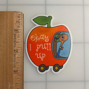 May include: A die-cut sticker shaped like an orange apple car with a green leaf on top, featuring a cartoon worm wearing a hat inside. The text 'okay i pull up' is printed in white. A wooden ruler shows the sticker is approximately 2.5 inches tall.