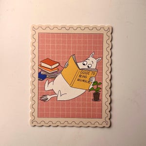 May include: A whimsical sticker featuring a white cartoon character reading a yellow book titled "Guide to Being Whimsical." The sticker has a pink grid background and a decorative border, with a small potted plant and a stack of books.