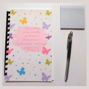 May include: A spiral-bound notebook with a butterfly design and the text "Rejoice always, pray continually, give thanks in all circumstances." A silver pen and a square of sticky notes are also present.