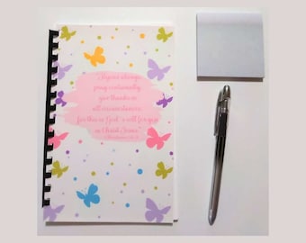 May include: A spiral-bound notebook with a butterfly design and the text "Rejoice always, pray continually, give thanks in all circumstances." A silver pen and a square of sticky notes are also present.