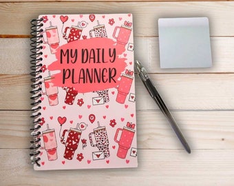 May include: Pink daily planner with a spiral binding and a cute design featuring pink and white mugs with hearts and flowers. The planner has the text "My Daily Planner" on the front cover.