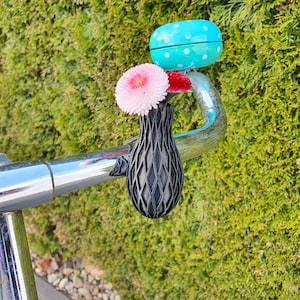Könnte beinhalten: Eine schwarze 3D-gedruckte Vase mit Gittermuster, die an einem verchromten Fahrradlenker befestigt ist. Die Vase enthält ein rosa und ein rotes Gänseblümchen. Eine helltürkise Fahrradklingel mit weißen Punkten ist neben der Vase am Lenker montiert.