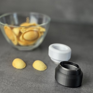 Könnte beinhalten: Ein ovaler Ausstechform aus schwarzem Metall und eine ovale Ausstechform aus weißem Kunststoff. Zwei kleine, hellbraune, ovale Kekse liegen auf der Oberfläche, während eine Glasschüssel mit ähnlichen ovalen Keksen dahinter platziert ist.