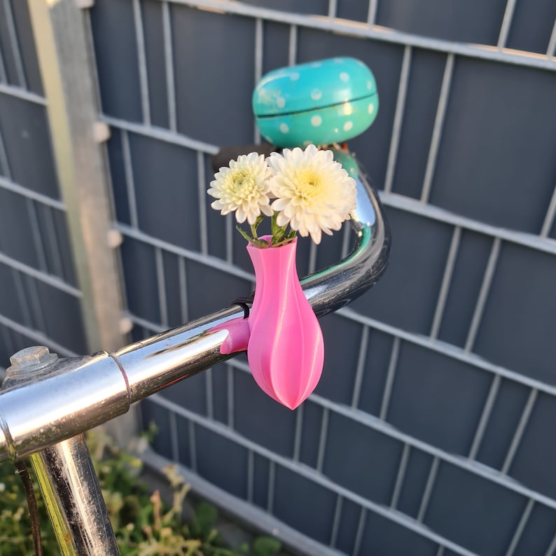 May include: A bright pink, bulbous, 3D-printed bicycle handlebar vase holding two white chrysanthemum flowers, positioned next to a turquoise bicycle bell with white polka dots on a metallic silver handlebar.