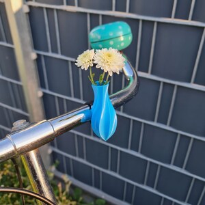 May include: A bright blue, spiraled, 3D-printed vase attached to a chrome bicycle handlebar. The vase holds two white chrysanthemum flowers. A turquoise bicycle bell with white polka dots is mounted on the handlebar behind the vase.