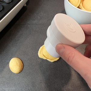 Könnte beinhalten: Ein weißes und beiges Küchengerät formt ein kleines, rundes, gelbes Backwaren. Im Hintergrund befindet sich eine Schüssel mit ähnlichen Backwaren. Das Gerät hat eine kreisförmige Oberseite mit dem Buchstaben "R".
