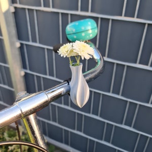 May include: A light gray, 3D-printed vase attached to a chrome bicycle handlebar. The vase holds three white chrysanthemum flowers. A bright turquoise bicycle bell with white polka dots is mounted on the handlebar behind the flower vase.