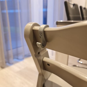 Könnte beinhalten: Ein hellgrauer S-förmiger Haken, der an der oberen Leiste eines Holzstuhls befestigt ist. Der Haken weist auf seiner Vorderseite eine kleine, eingravierte Herz-Kontur auf. Der Stuhl hat eine glatte, helle Oberfläche und eine geschwungene Rückenlehne.