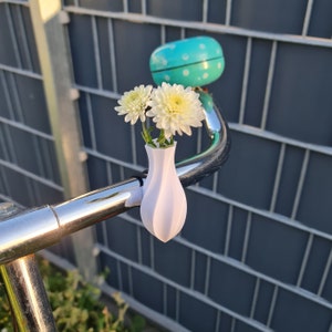 May include: A white, 3D-printed, teardrop-shaped vase attached to a chrome bicycle handlebar. The vase holds two white chrysanthemum flowers. A bright turquoise bicycle bell with white polka dots is mounted on the handlebar behind the vase.