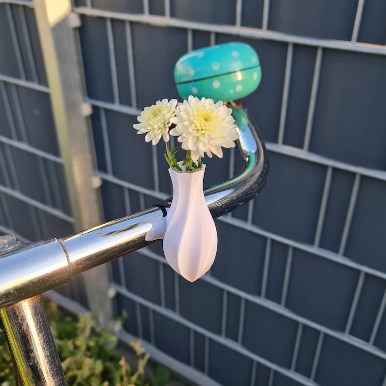 May include: A white, 3D-printed, teardrop-shaped vase attached to a chrome bicycle handlebar. The vase holds two white chrysanthemum flowers. A bright turquoise bicycle bell with white polka dots is mounted on the handlebar behind the vase.