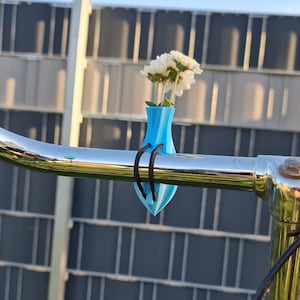 May include: A bright blue, teardrop-shaped vase attached to a shiny chrome bicycle handlebar with a black elastic cord. The vase holds a small bouquet of white flowers with yellow centers, positioned upright against the curved metal bar.