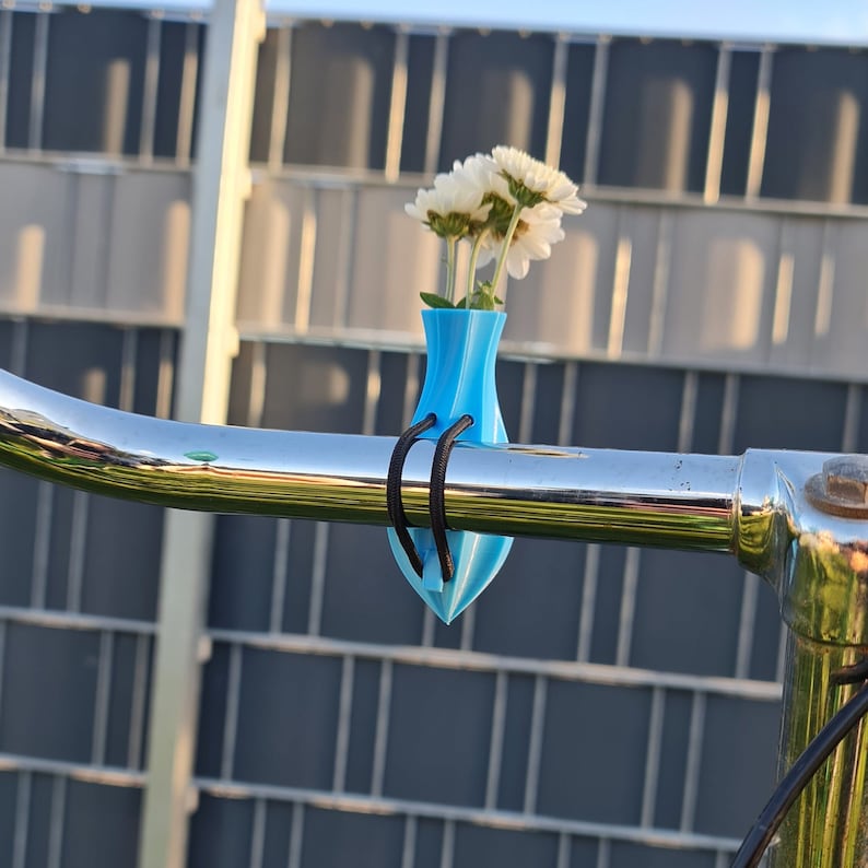 May include: A bright blue, teardrop-shaped vase attached to a shiny chrome bicycle handlebar with a black elastic cord. The vase holds a small bouquet of white flowers with yellow centers, positioned upright against the curved metal bar.