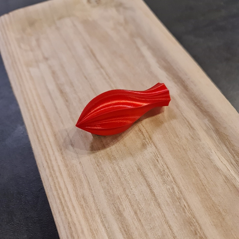 May include: A bright red, three-dimensional, teardrop-shaped object with a series of vertical, curved ridges running along its length. The object has a pointed tip at one end and a slightly wider, flat-edged opening at the opposite end.