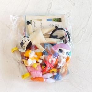 May include: A clear plastic bag containing a collection of assorted small toys, including a cream-colored fabric doll, a small orange and white fox figure, a clown figurine, a doll with purple hair, and various miniature plastic accessories.