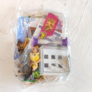 Op de afbeelding: Een doorzichtige plastic zak met een verzameling kleine voorwerpen, waaronder een paarse zandloper, een witte plastic doos met zwarte stippen, diverse witte dobbelstenen, een klein geel figuurtje en kleurrijke papieren kaarten met de tekst NERDS IV.
