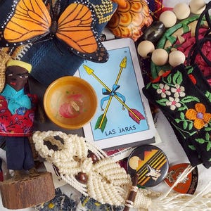 Op de afbeelding: Een verzameling voorwerpen waaronder een monarchvlinderornament, een kleine pop op een houten voet, een tarotkaart met het label LAS JARAS, een gele houten kom, een zwarte geborduurde stoffen strook, een macraméstuk en diverse kralen.