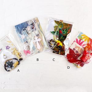 Op de afbeelding: Vier doorzichtige plastic zakjes met de labels A, B, C en D, elk met diverse kleine voorwerpen. Zakje A bevat een zwarte ster en gouden bedels. Zakje B bevat een lichtblauw kruis en roze dierenfiguren. Zakje C bevat groene en gele kralen. Zakje D toont een afbeelding van een kat met rode stof.